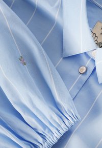 Světlomodrá košile s tenkými bílými vertikálními pruhy, zapínaný límec, nařasený elastický manžet a malý vyšívaný fialový květinový detail.