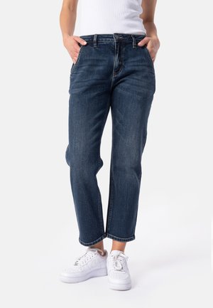 Person iført mørkeblå jeans og hvide sneakers, stående med hænderne i lommerne mod en ensfarvet baggrund.