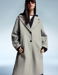 Femeie îmbrăcată cu un sacou oversized de culoare gri deschis, cu butoni, peste un hanorac negru, pe un fundal simplu de gri-albăstrui.