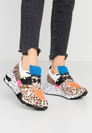 Füße tragen klobige Sneakers mit gemischten Animal-Prints, orange und blaue Akzente, schwarze Schnürsenkel und schwarze Jeans mit ausgefransten Säumen.