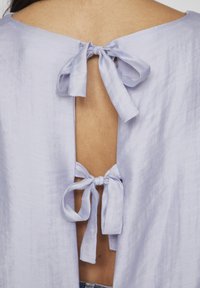 Bluză lavandă deschisă cu un decupaj la spate și două fundițe. Textură netedă cu un luciu subtil. Design casual, cu o croială relaxată.