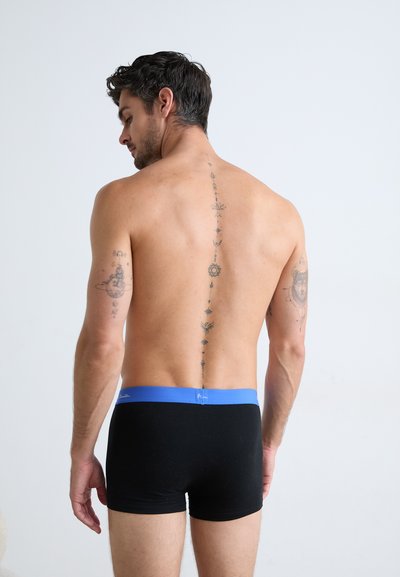 Boxers noirs avec une ceinture bleue. Le modèle a un tatouage courant verticalement le long du dos, mettant en avant des motifs complexes.
