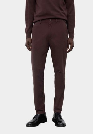 Mand iført mørkebrune slim fit bukser med matchende sweater og sorte polerede finere sko på en ensfarvet lys baggrund.