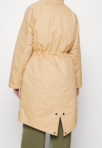 Light beige long coat made of smooth fabric, featuring a cinched waist, long sleeves, and a back vent with button accents.