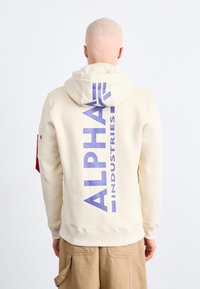 Sweat à capuche crème avec un grand imprimé violet "ALPHA INDUSTRIES" dans le dos, doté d'une poche kangourou et d'une capuche réglable. Matière en coton.