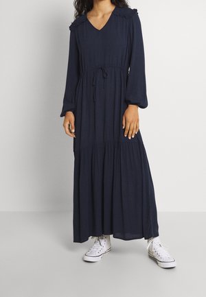 Kobieta w długiej granatowej sukience z długimi rękawami i białych wysokich trampkach, stojąca na tle jednolitego jasnego tła.