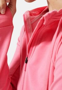 Chaqueta rosa con cremallera, puños y cuello acanalados, con una textura suave y una pequeña etiqueta con el logo dentro del escote. Tirador de cremallera en negro.