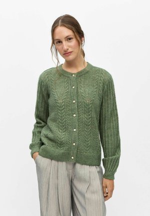 Femme portant un cardigan en maille torsadée vert avec des boutons en perle et un pantalon plissé gris clair, debout avec une main dans la poche.