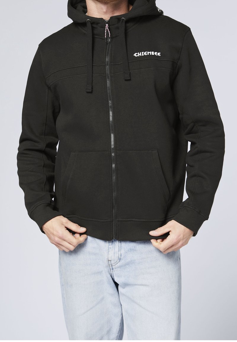 Homme portant un sweat à capuche noir zippé avec poches avant et logo blanc "Chiemsee", associé à un jean bleu clair, tenant les bords du sweat.