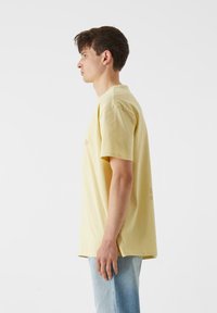 Geltonas medvilninis marškinėliai trumpomis rankovėmis, su apvaliu kaklu ir laisvu pritaikymu, demonstruojami profilio modelio. Šviesiai mėlyni džinsai papildo derinį.