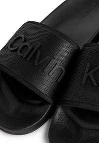 Mustad must toastega silmadega mustad kummist sandaalid, millel on kõrgendatud "Calvin" ja "Klein" brändimine; sile tald õrna tera viimistlusega.