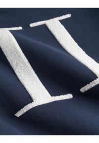 Tejido azul marino con letras blancas texturizadas. El material es suave, con una superficie lisa y un bordado esponjoso, que destaca el diseño de las letras.