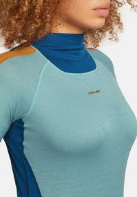 Donna che indossa un top a maniche lunghe in lana merino blu e turchese con toppe arancioni sulle spalle e un collo alto.