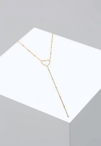 Guld halskæde med et hjerteformet vedhæng, der har en delikat kæde med små perler, hængende fra en hvid geometrisk display.