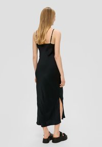 Schwarzes Slip-Kleid mit dünnen Spaghettiträgern, seitlichen Schlitzen und einer glatten, glänzenden Textur. Kombiniert mit schwarzen Plateausandalen.