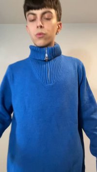 Jeune personne aux cheveux courts et foncés portant un pull bleu zippé, debout contre un mur clair uni.