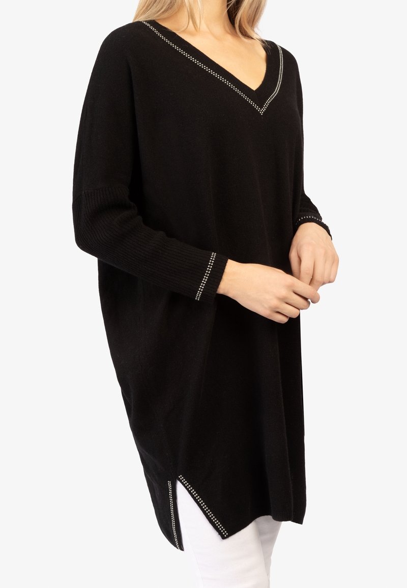 Tunique pull noire à manches longues avec col en V et petits clous décoratifs sur le col, les poignets et les fentes latérales, portée sur un pantalon blanc.