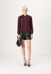 Burgunderfarget zip-up jakke med lange ermer, kombinert med svarte skinnshorts. Modellen holder en svart håndveske og har på seg svarte høyhælte sandaler.