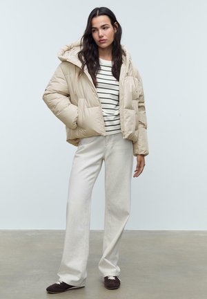 Doudoune beige avec une capuche, poches frontales, associée à une chemise rayée blanche et un pantalon ample crème. Des chaussures marron foncé complètent l'ensemble.