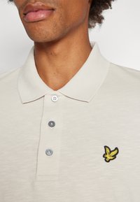 Bežna polo majica s tremi srebrnimi gumbi, z vezenim logotipom ptice v črni in rumeni barvi na levem prsnem delu.