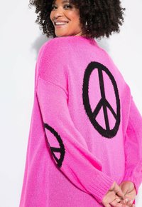 Růžový velký svetr s dlouhými rukávy, zdobený velkými černými vzory peace sign na obou stranách a texturovanou pletenou látkou.