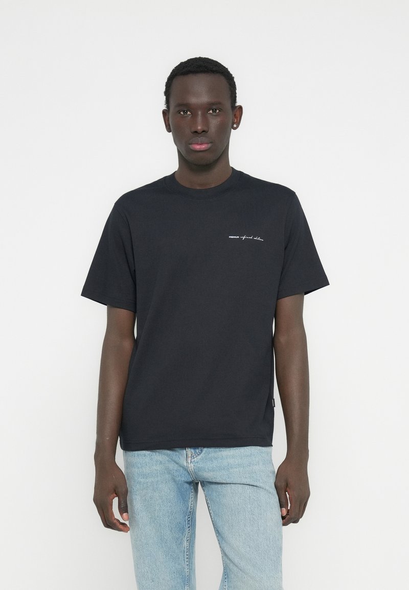 Μαύρο βαμβακερό t-shirt με στρογγυλή λαιμόκοψη, κοντά μανίκια και μικρή λευκή λεπτομέρεια κειμένου στο μπροστινό μέρος. Φορεμένο με ανοιχτόχρωμα μπλε τζιν.