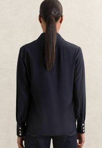 Navy button-up shirt made of smooth fabric, featuring long sleeves with button accents and a structured collar. Back view shown.