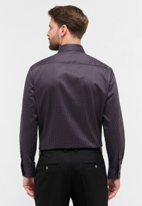 Paarse blouse met een subtiele stippenpatroon, lange mouwen en een klassieke kraag. Draagt samen met zwarte broek met zijzakken.