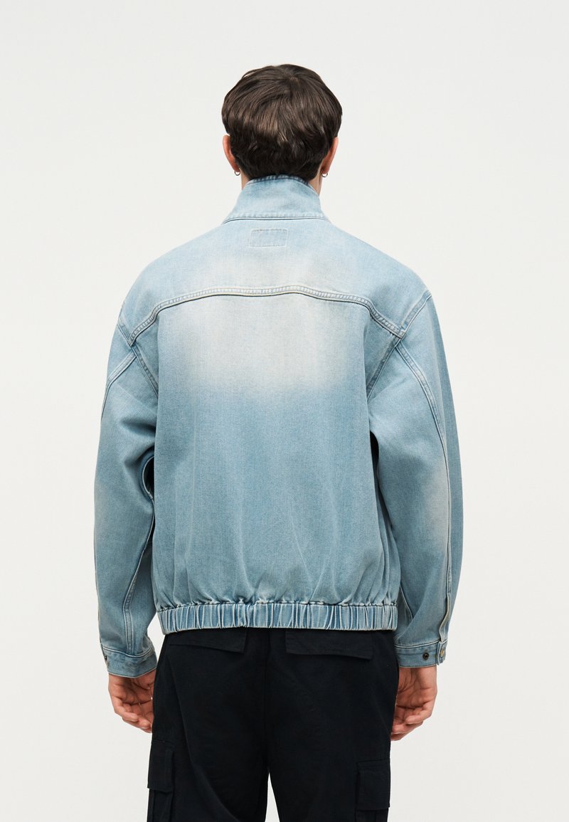 Uomo con giacca di denim azzurro chiaro e pantaloni neri, visto di spalle su uno sfondo bianco tinta unita.