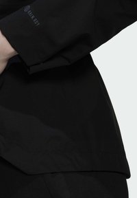 Zwarte jas met een gladde textuur, met een licht getailleerde vorm en een logo op de mouw. De naaddetails zijn subtiel en verfijnd.