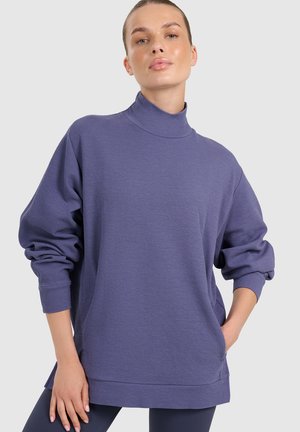 Kobieta ubrana w luźny fioletowy golf z długimi rękawami i granatowe legginsy, stoi z jedną ręką w kieszeni.