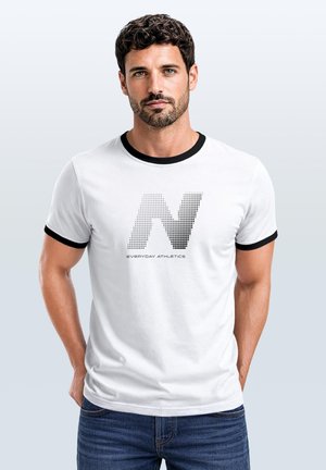Mann mit kurzen lockigen Haaren, der ein weißes T-Shirt mit schwarzem Besatz und großem gepunktetem "N Everyday Athletics" Logo trägt, stehend vor hellem Hintergrund.