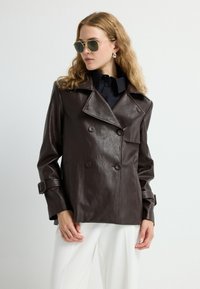 Giacca doppiopetto in finta pelle marrone con grandi revers, tasche laterali e dettagli con fibbia. Il modello indossa occhiali da sole verdi e una maglietta nera.