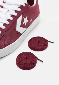 Burgundy suede sneaker with white textile laces and a star logo. Two rolled burgundy replacement laces are positioned nearby.