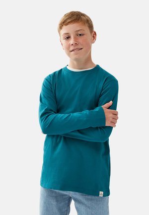 Teal langærmet skjorte i bomuld med rund halsudskæring; har et lille mærke ved sømmen og bæres med lyseblå jeans.