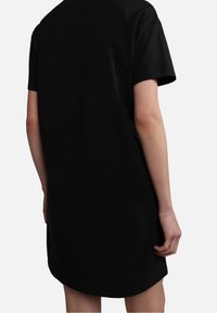 Person stående med ryggen til kameraet, iført en kortærmet sort kjole foran en plain hvid baggrund.