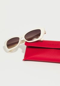 Gafas de sol blancas de gran tamaño con lentes gradientes oscuros junto a un estuche de cuero rojo texturizado, grabado con el logo "GUESS" en la parte frontal.