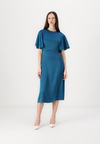 Robe en satin turquoise avec des manches volantes, longueur mi-mollet, taille cintrée, texture lisse, associée à des talons hauts de couleur claire.