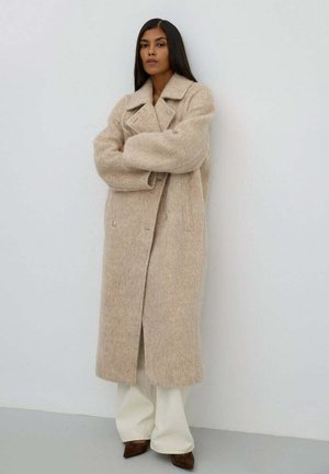 Béžový vlněný kabát s klopovým límcem, dvouřadým zapínáním a bočními kapsami, nošený přes bílé kalhoty. Hladká textura.