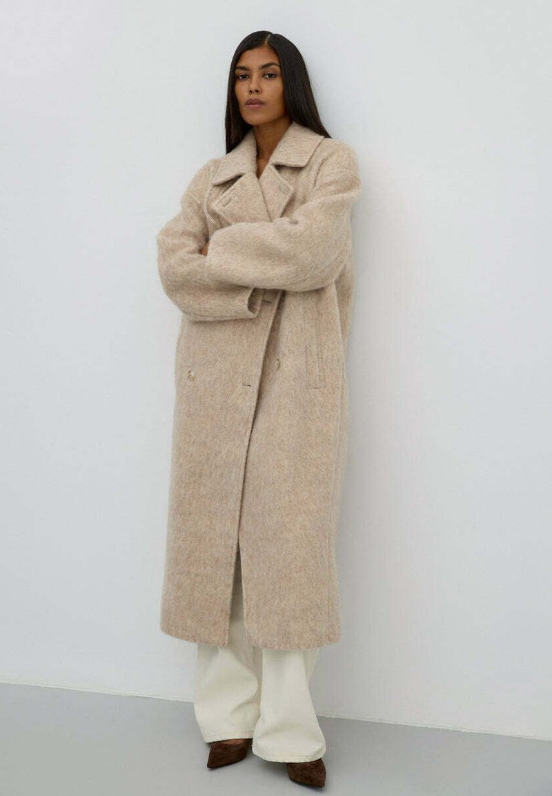 Béžový vlněný kabát s klopovým límcem, dvouřadým zapínáním a bočními kapsami, nošený přes bílé kalhoty. Hladká textura.