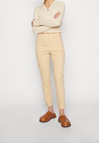 Personne portant un cardigan cintré beige à boutons, un pantalon taille haute beige et des chaussures brunes à enfiler, debout les bras croisés.