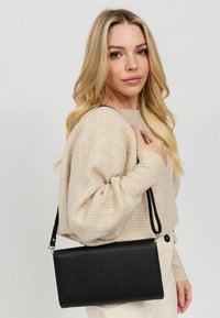 Zwarte neplederen schoudertas met een rechthoekige vorm, gouden hardware en een afneembare schouderband, gedragen over een beige gebreide trui.