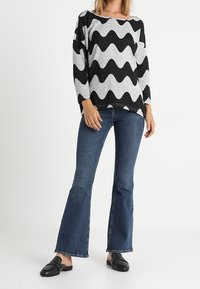 Schwarzer und grauer Pullover mit wellenförmigem Muster und langen Ärmeln, kombiniert mit ausgestellt geschnittenen blauen Jeans und schwarzen Slippers mit Nietenverzierung.