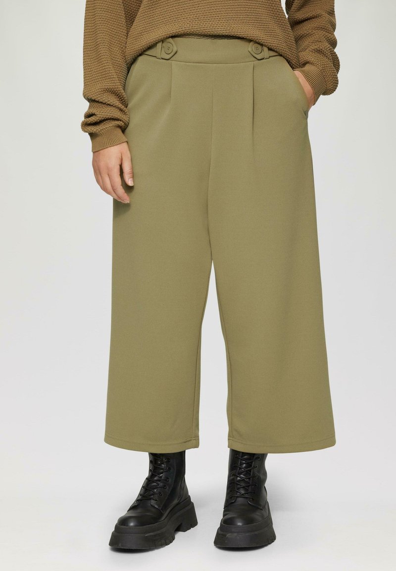 Olijfgroene wijde broek met plooien en knoopdetails bij de taille, gecombineerd met zwarte veterschoenen met plateauzolen. Gladde textuur.