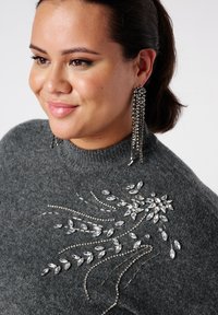 Grijze gebreide trui met ingewikkelde kristallen versieringen, met een bloemmotief. Lange, hangende juwelenoorbellen maken de look compleet.