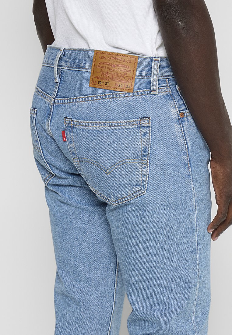 Persona que lleva puestos jeans Levi's 501 de color azul claro con el bolsillo trasero visible, etiqueta roja y parche de cuero marrón de la marca en la cinturilla.