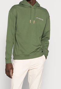 Muž v olivově zelené mikině s tkaničkami a bílých kalhotách, s jednou rukou v kapse, stojící před jednoduchým světlým pozadím.