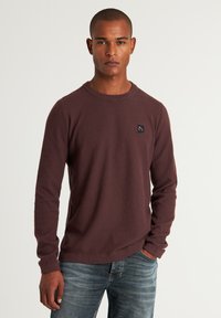 Sweatshirt bordo cu mâneci lungi din material texturat, decolteu rotund, manșete ribbate și o mică etichetă logo neagră pe piept.