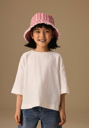 Smaidīgs bērns rozā svītrains cepurē, vaļīgā baltā īsroku krekliņā un zilās džinsās uz vienkārša bēšā fona.