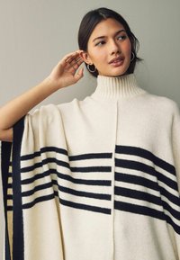 Cream-colored poncho with a high ribbed collar, featuring black horizontal stripes, soft texture, and open sides for ease of movement.